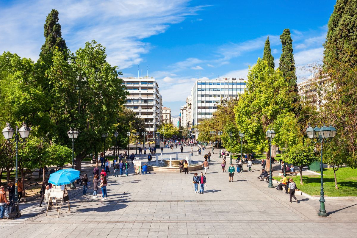 A spacious Syntagma square in Athens lined with trees and lampposts, with people walking and vendors under blue umbrellas.